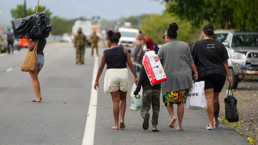 a family walks down the side of a closed highway carrying bags of belongings