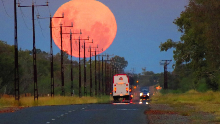 A red full moon rises over vehicles driving along a road south of Alice Springs