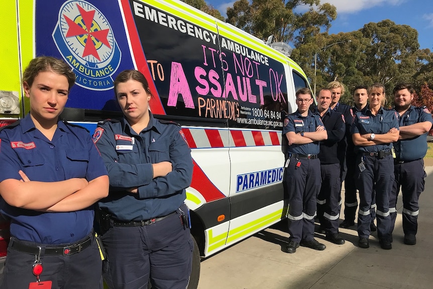 Eight paramedics with arms crossed stand beside an ambulance which has 'it's not ok to assault paramedics' written on it