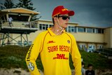 Spyros wearing a bright yellow surf rescue shirt and a red cap, standing in front of a surf lifesaving club and looking around.