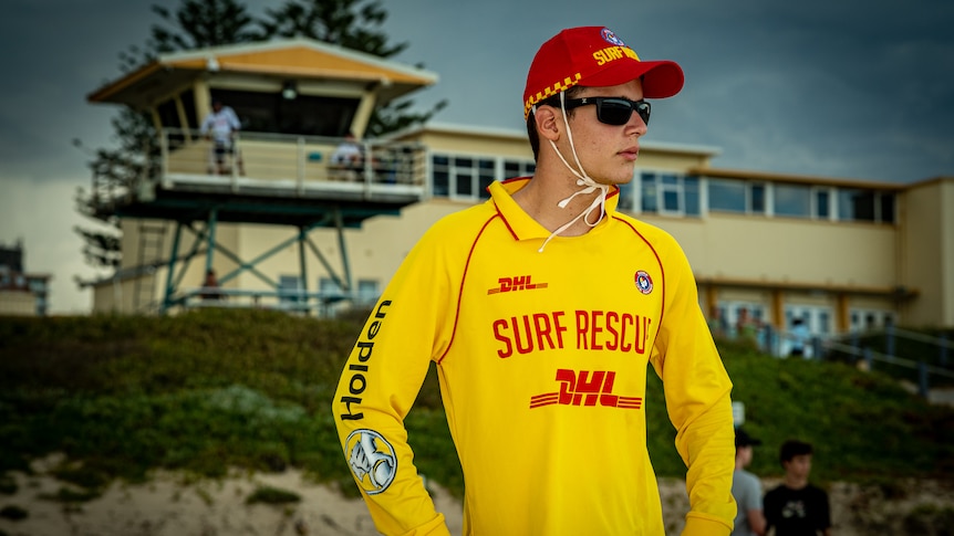 Spyros wearing a bright yellow surf rescue shirt and a red cap, standing in front of a surf lifesaving club and looking around.