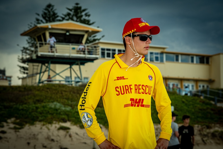 Spyros wearing a bright yellow surf rescue shirt and a red cap, standing in front of a surf lifesaving club and looking around.