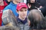 A teenager in a MAGA hat stares at a native American protester as he plays a drum as a crowd also wearing MAGA hats watches on