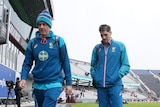 Two men walk off a cricket field looking cold and wet.