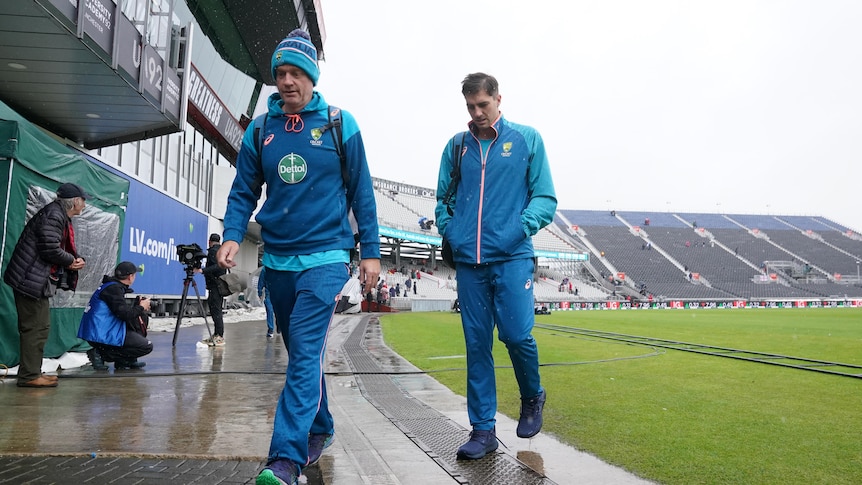 Two men walk off a cricket field looking cold and wet.