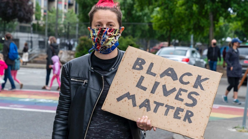 Woman wearing mask, black leather jacket and black top with pink and blonde hair holds Black Lives Matter sign