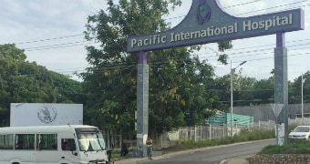 A hospital sign reads "Pacific International Hospital" on a cloudy day with a van out front and two men stood by sign.