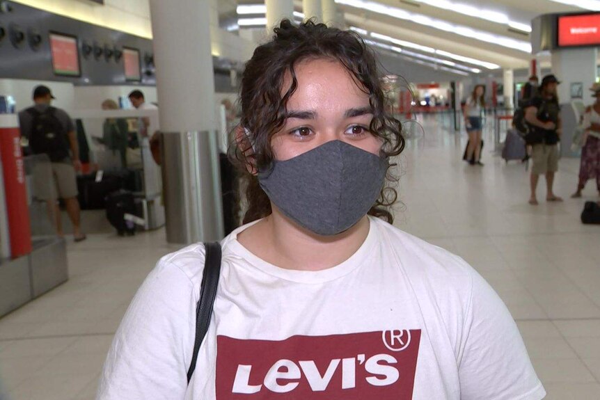 A woman wearing a grey face mask stands in Perth Airport with passengers in the background.