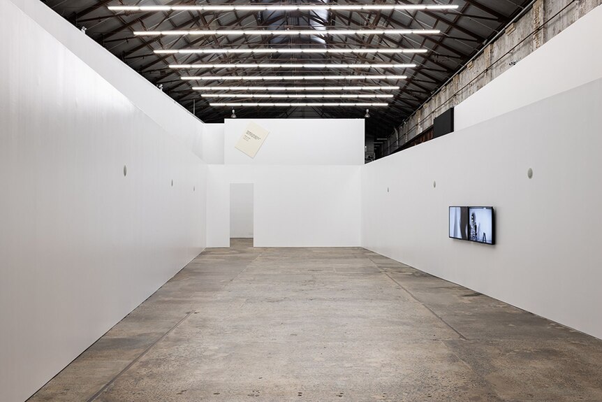 White fluorescent lights hang from pitched tin roof above large white gallery space with concrete floor and single tv installed.