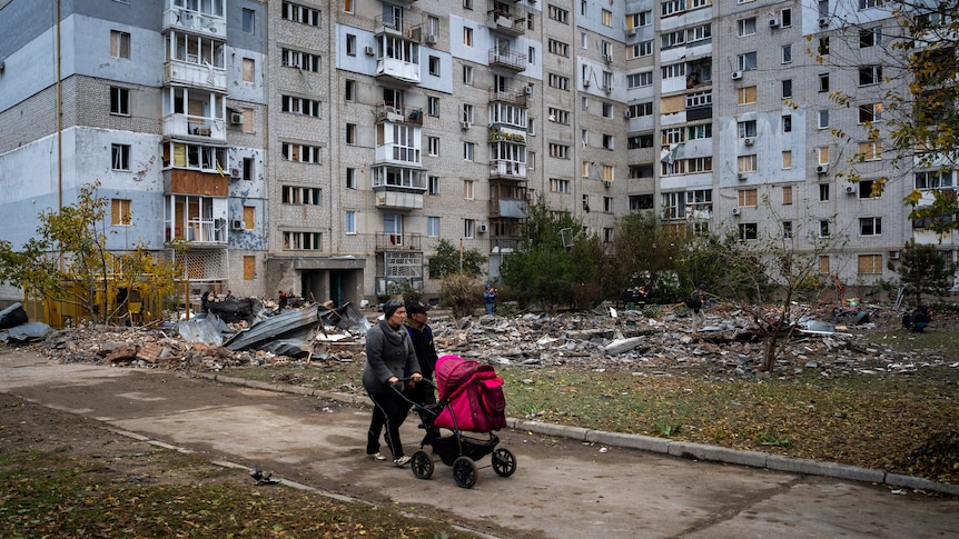 A couple pushing a baby stroller walks past a building damaged by a Russian missile.