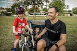 Kid on bike, kid in running pram, and dad in a park.
