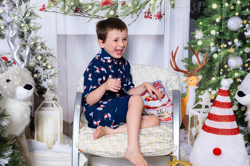 A boy in Christmas-themed clothes sits between Christmas trees and decorations, laughing.