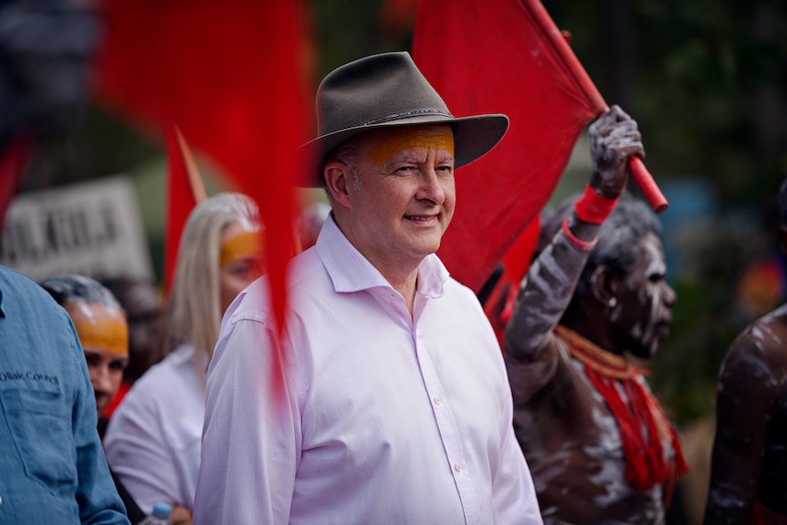 Anthonty Albanese wearing a pink shirt and a wide brimmed hat surrounded by red flags and people in body paint.