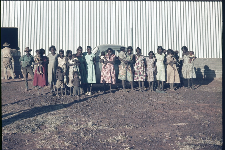 A colour photograph from 1962 depicting a group of Aboriginal women and children posing for a photo in front of a large shed.