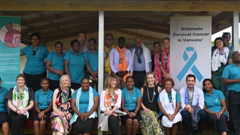 A group of staff wearing blue shirts pose for a photo near a banner saying 'Eliminate cervical cancer in Vanuatu'.