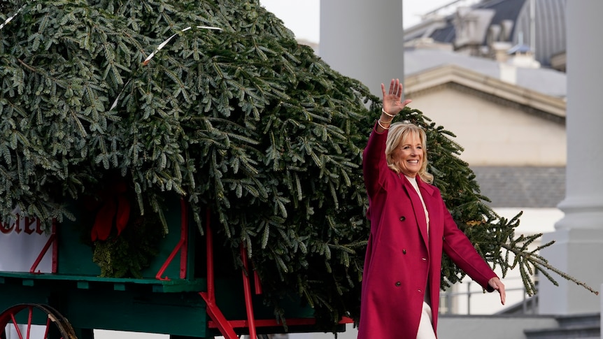 A woman wearing a pink coat waves in front of a huge Christmas tree.