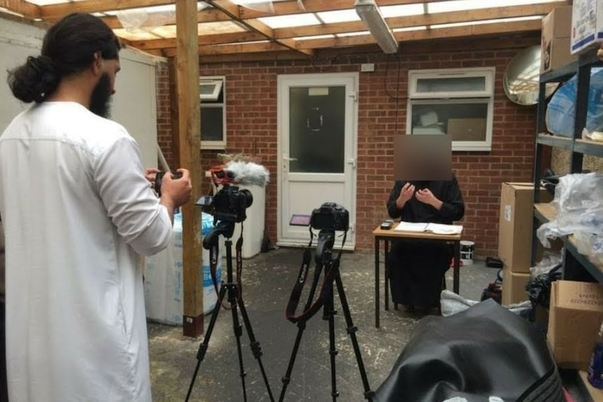 A bearded man operates a camera while another unidentified man sits at a table.