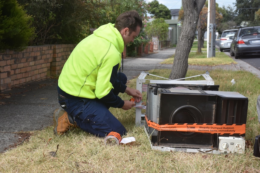 A man in a yellow hi-vis hoodie and jeans kneels beside a dumped air conditioning unit on a grassy footpath.