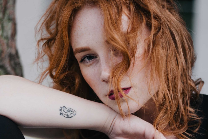Woman with red hair showing off a small fine line tattoo on her arm for a story about what you need to know before getting one.