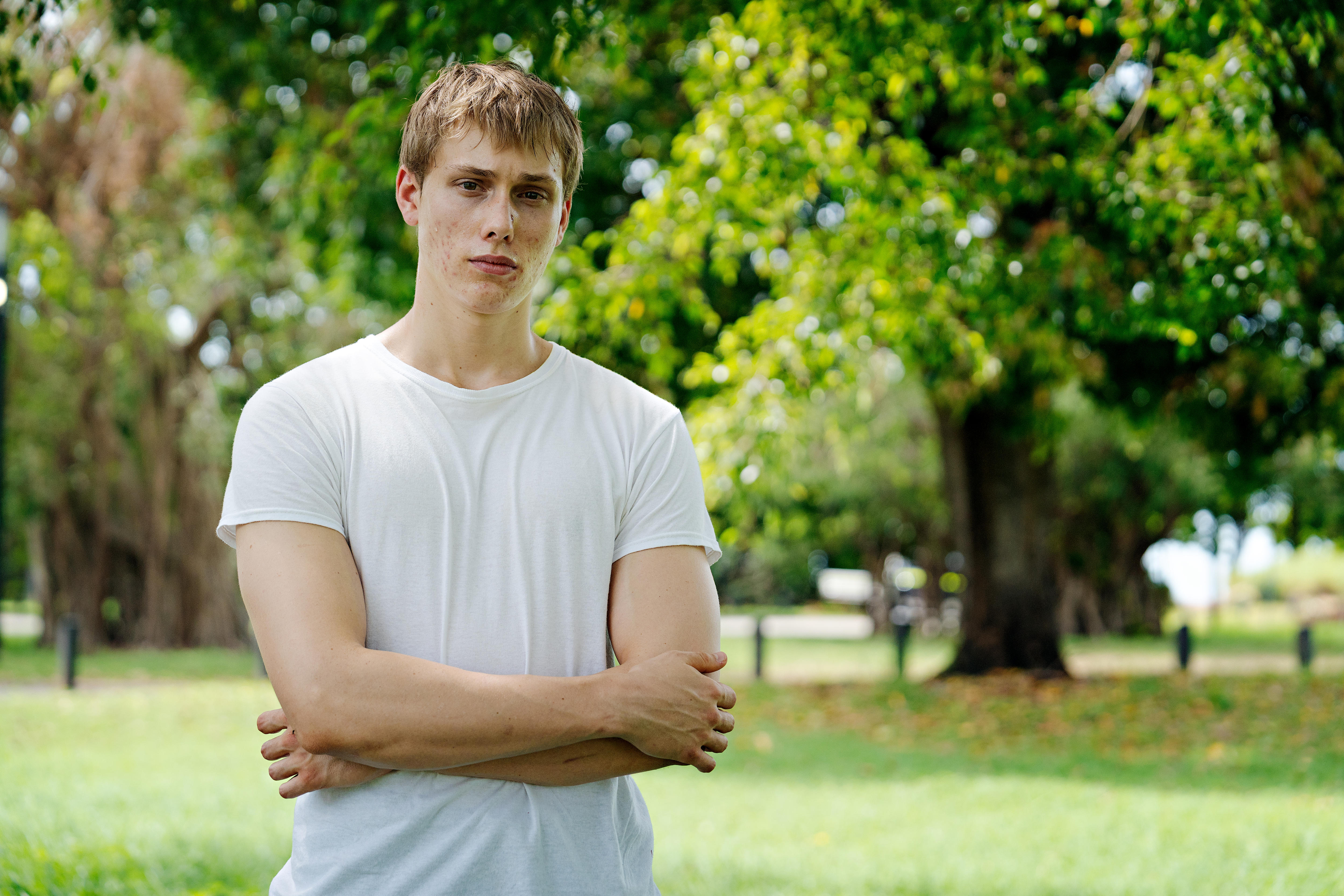 Um homem vestindo uma camisa branca com os braços cruzados olha seriamente para a câmera.