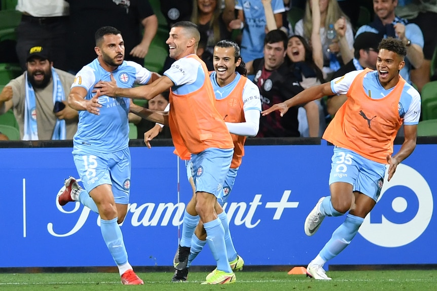 Un jugador de fútbol de pelo oscuro vestido de azul celebra un gol rodeado de tres compañeros con baberos naranjas