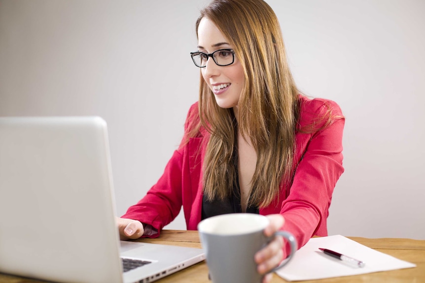 A woman in glasses and a red blazer sits in front of a laptop and a cup of coffee