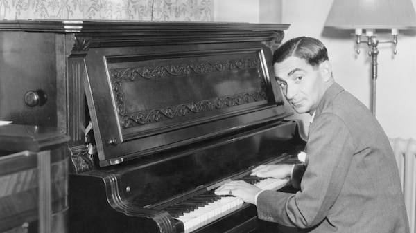 A black and white photograph of a man in a suit sitting at an upright piano.