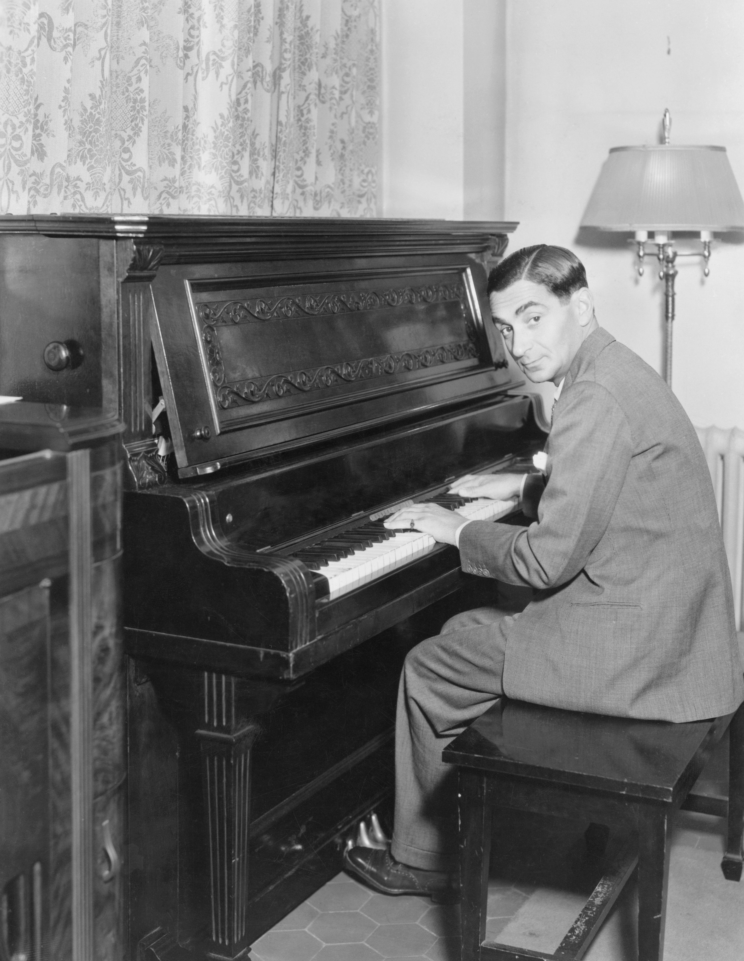 A black and white photograph of a man in a suit sitting at an upright piano.