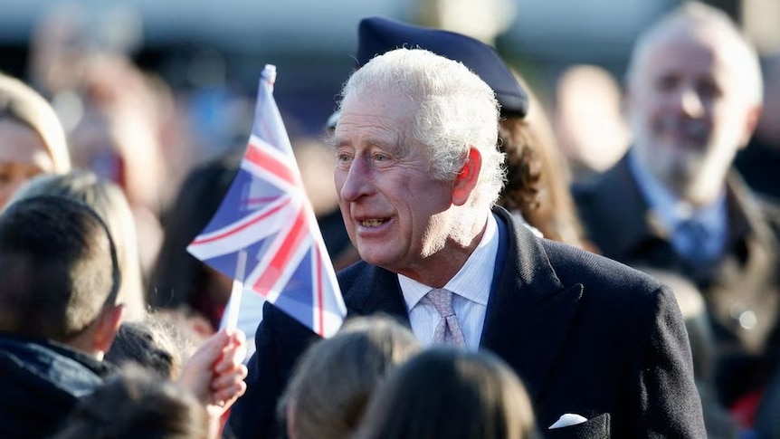 King Charles III smiling in a crowd of people waving small United Kingdom flags 