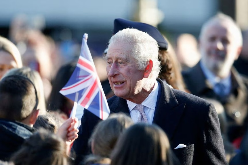 King Charles III smiling in a crowd of people waving small United Kingdom flags 