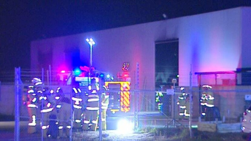 Firefighters and fire trucks stand outside a warehouse in Port Kennedy at night with black marks on the walls of the buidling.
