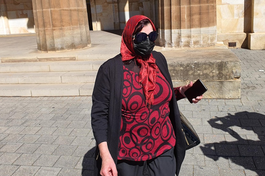 A woman wearing a red head scarf and shirt, and a black jacket and face mask, and sunglasses outside a sandstone building