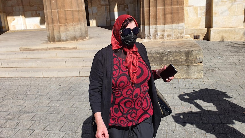 A woman wearing a red head scarf and shirt, and a black jacket and face mask, and sunglasses outside a sandstone building