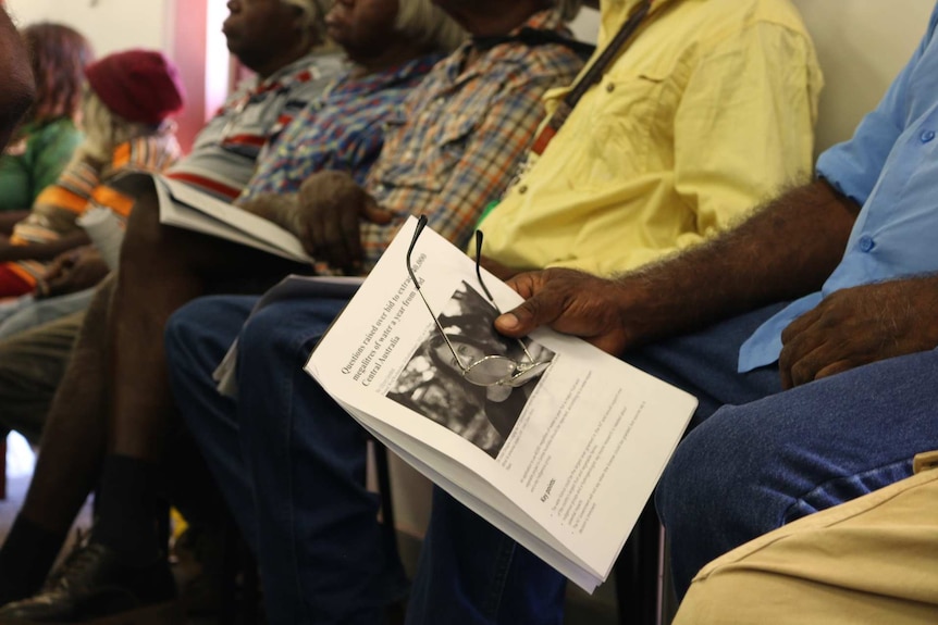 A group of Indigenous people sit in a room. One of them holds a pair of glasses and a document.