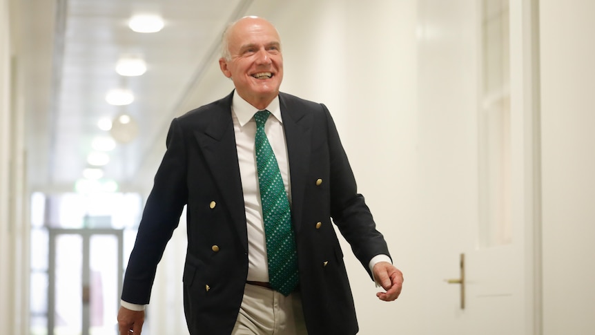 Abetz smiles while wearing a dark suit jacket, tan pants and green tie.