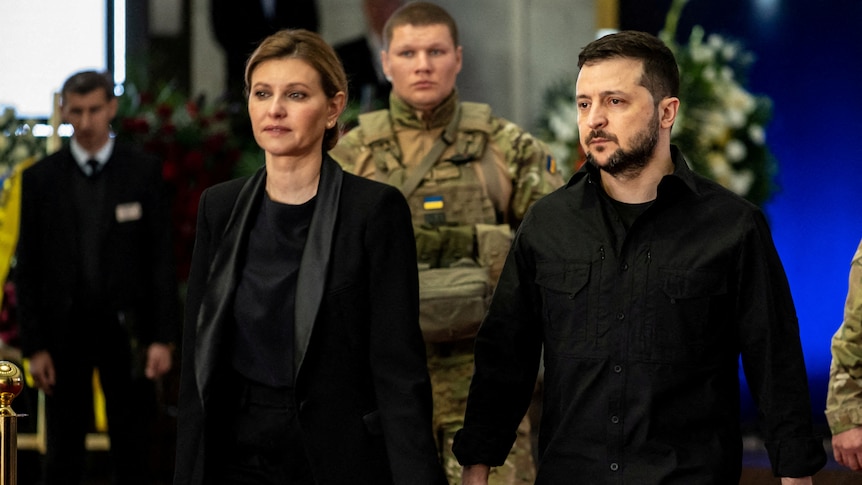 Volodymyr Zelenskiy walks and holds the hands of his wife with soldiers behind him in sombre ceremony