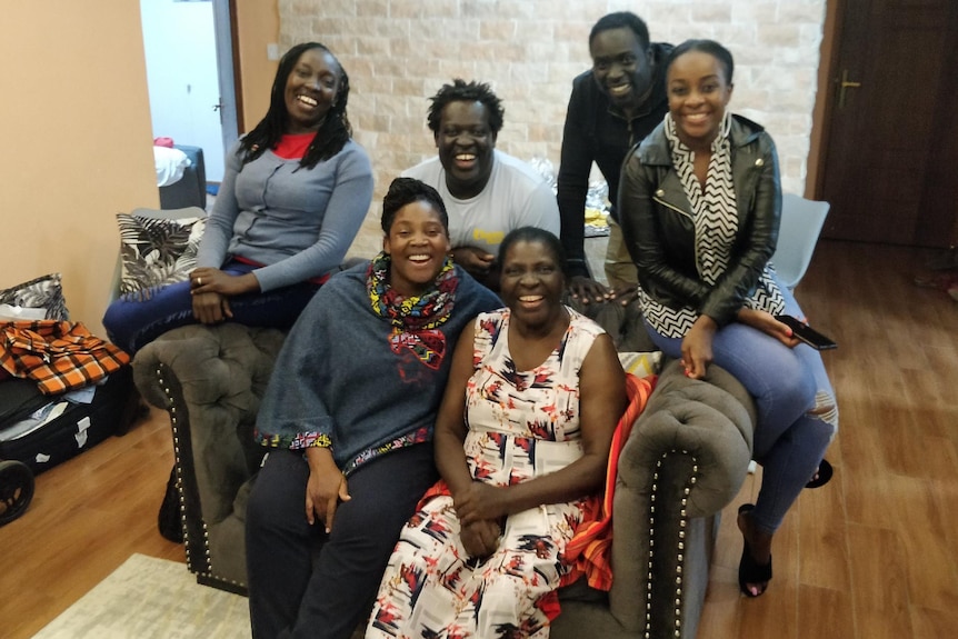 Four women and 2 men sitting and standing around a sofa close together and laughing in a home setting. 