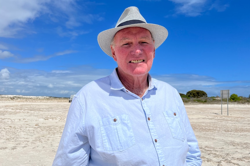 Mann steht in weißen Sanddünen mit Hut und hellblauem Hemd und lächelt