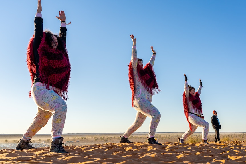 Three women wear onsie pyjamas and red shawls doing yoga in the desert.