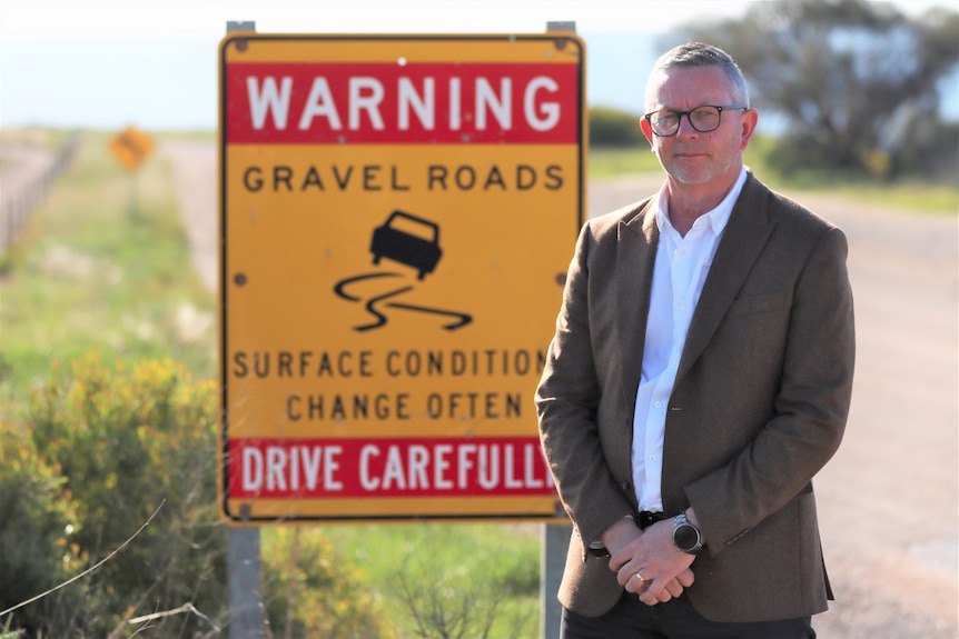 A grey-haired man wears a white shirt and brown jacket in front of a yellow sign.