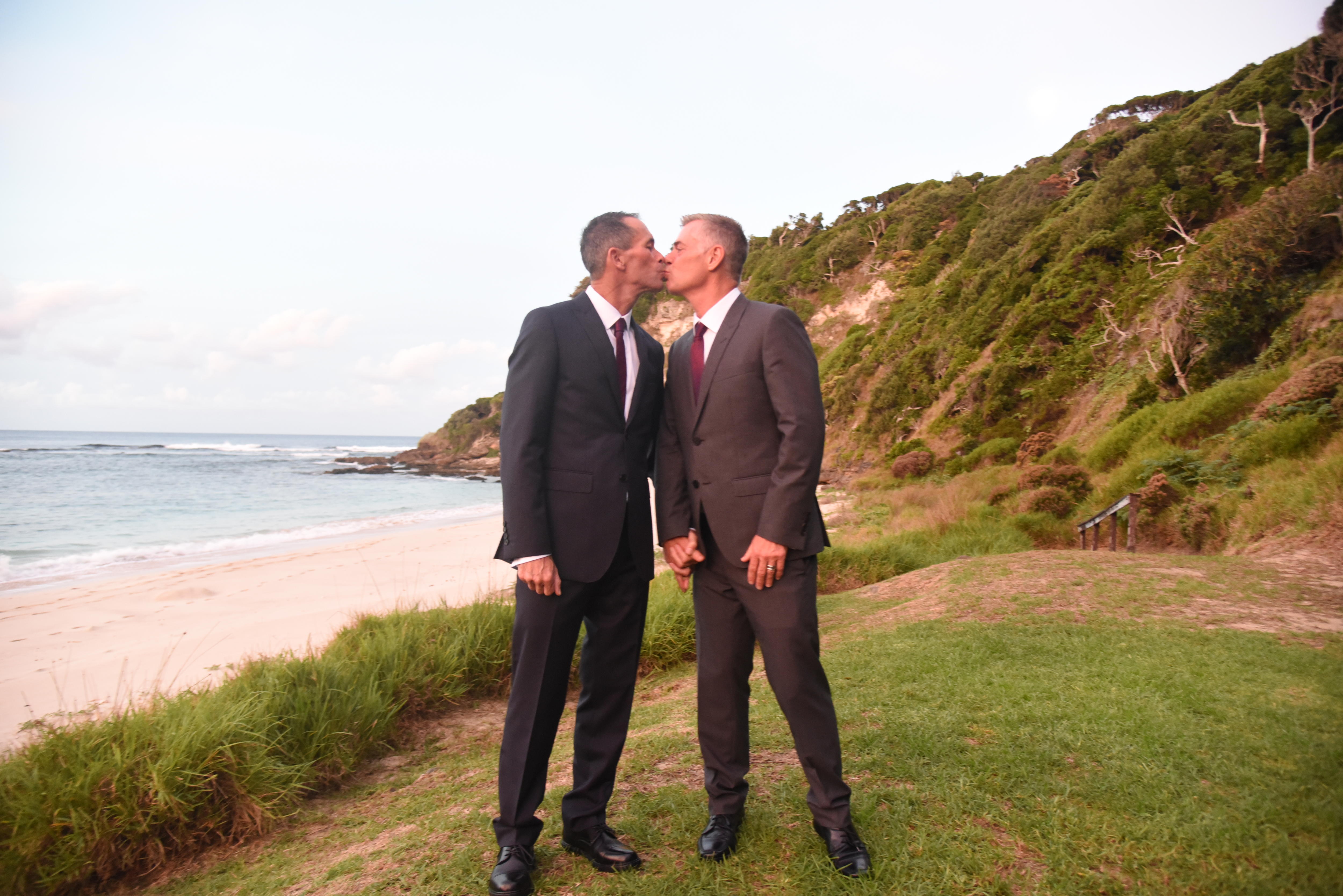 Two men wearing suits stand together on an island, holding hands and kissing each other on their wedding day.