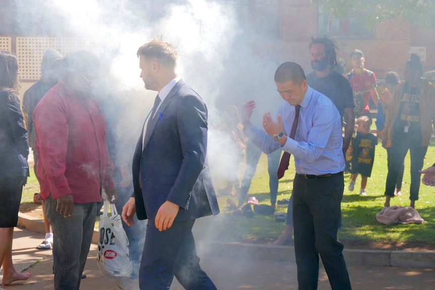 A man with a blue shirt walks through smoke alongside a man with a blue suit. 