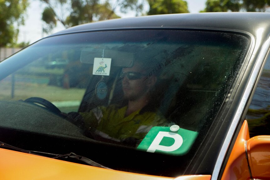 A man behind the wheel with glasses on and a green p plate in the car window.