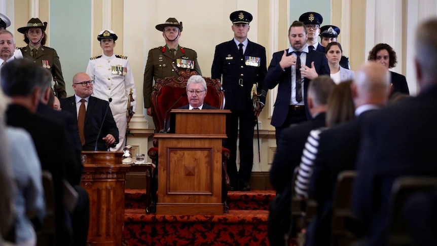 Man in official chair with members of the armed forces behind him and others standing nearby