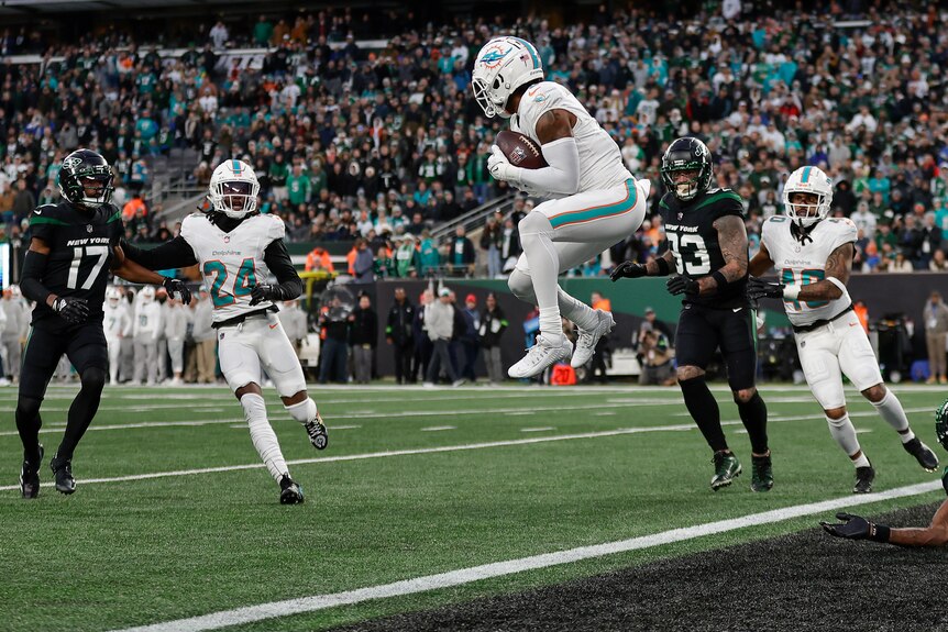 A Miami NFL defender leaps in the air to catch a ball as his teammates and rival Jets players look on.