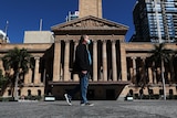 a man with a face mask in a suit jacket and jeans walks past Brisbane's city hall in daytime.