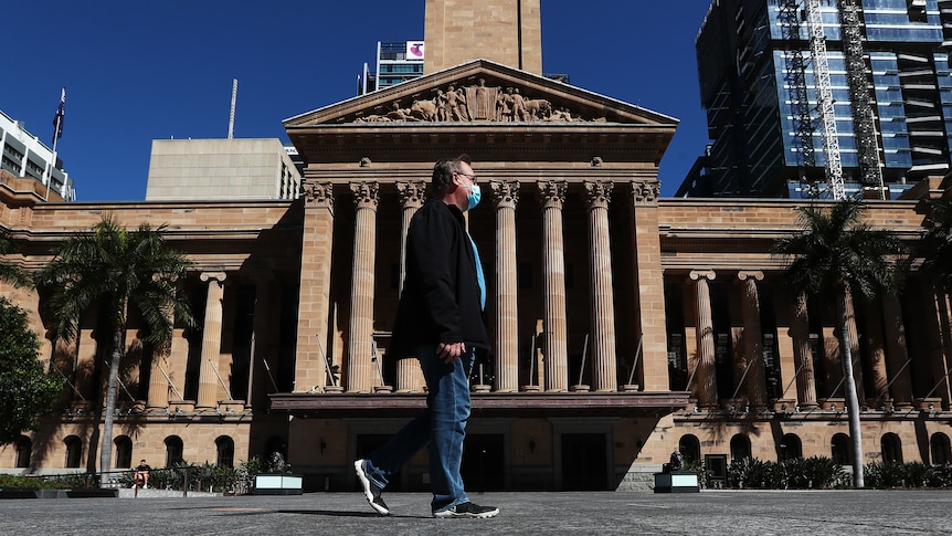 a man with a face mask in a suit jacket and jeans walks past Brisbane's city hall in daytime.