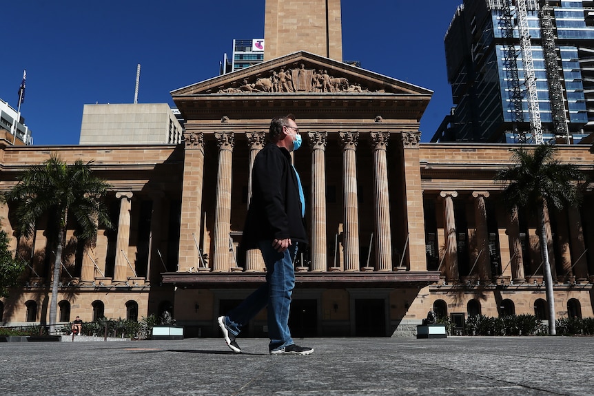 a man with a face mask in a suit jacket and jeans walks past Brisbane's city hall in daytime.