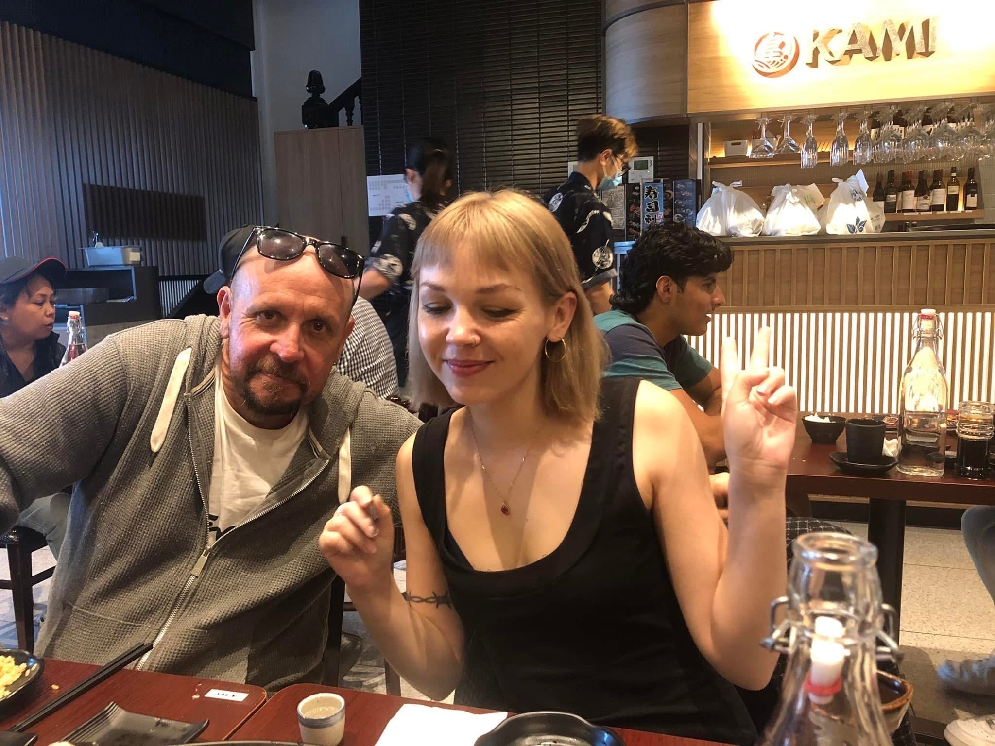 Un hombre que lleva una sudadera con capucha gris se sienta en la mesa de un restaurante junto a una mujer joven con cabello rubio y un top negro. 