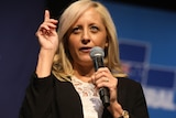 Melissa McIntosh gestures with her hand, pointing up, while holding a microphone. Behind her is a Liberal party logo.